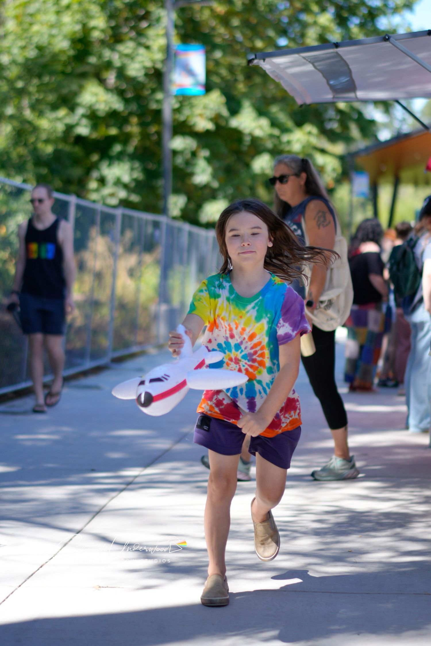 Smiling child in a tie-dye shirt runs down a sunny street fair path holding a foam toy airplane while crowds gather behind.