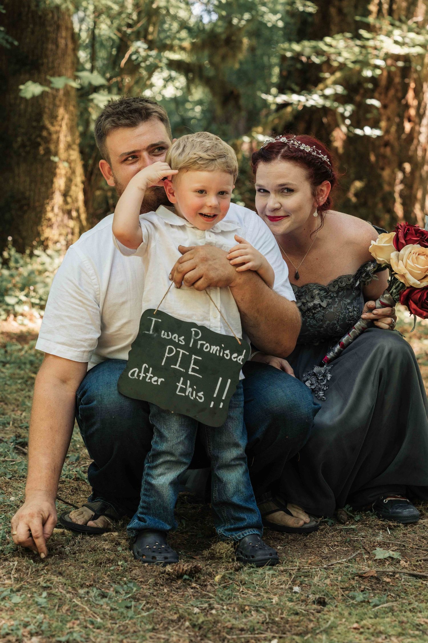 A couple kneels with their toddler son wearing a chalkboard sign reading I was promised pie after this, in a forest wedding portrait.