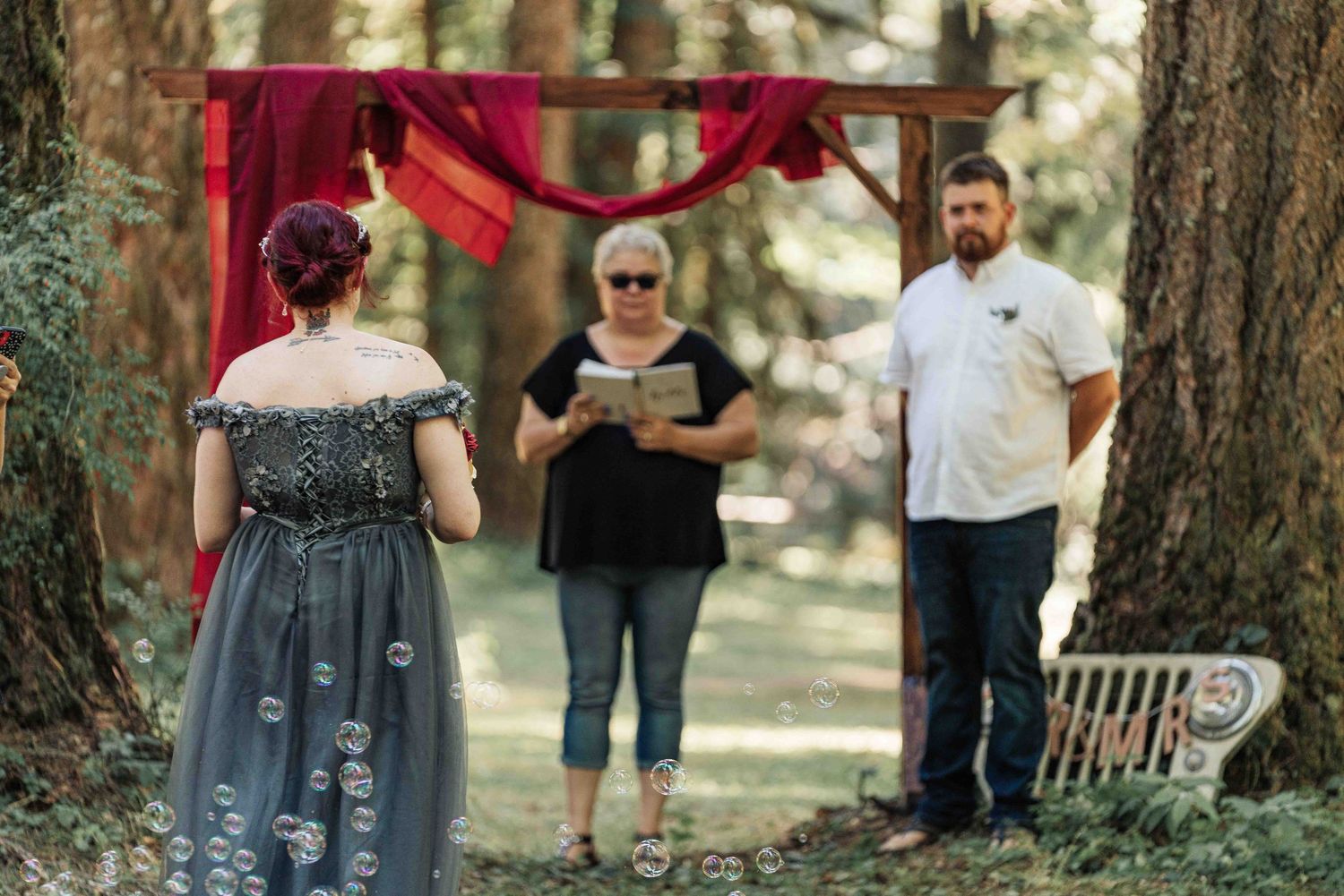 A bride in a dark gown with bubbles floating around her listens to the officiant as the groom looks on in the forest.