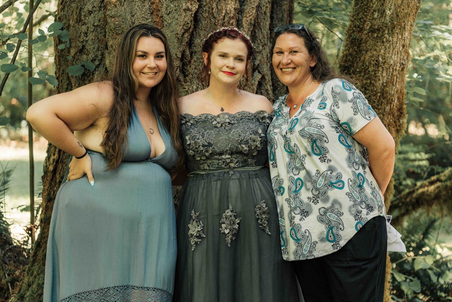 Three women pose together beside a large mossy tree, the bride in a gray gown flanked by two smiling wedding guests.
