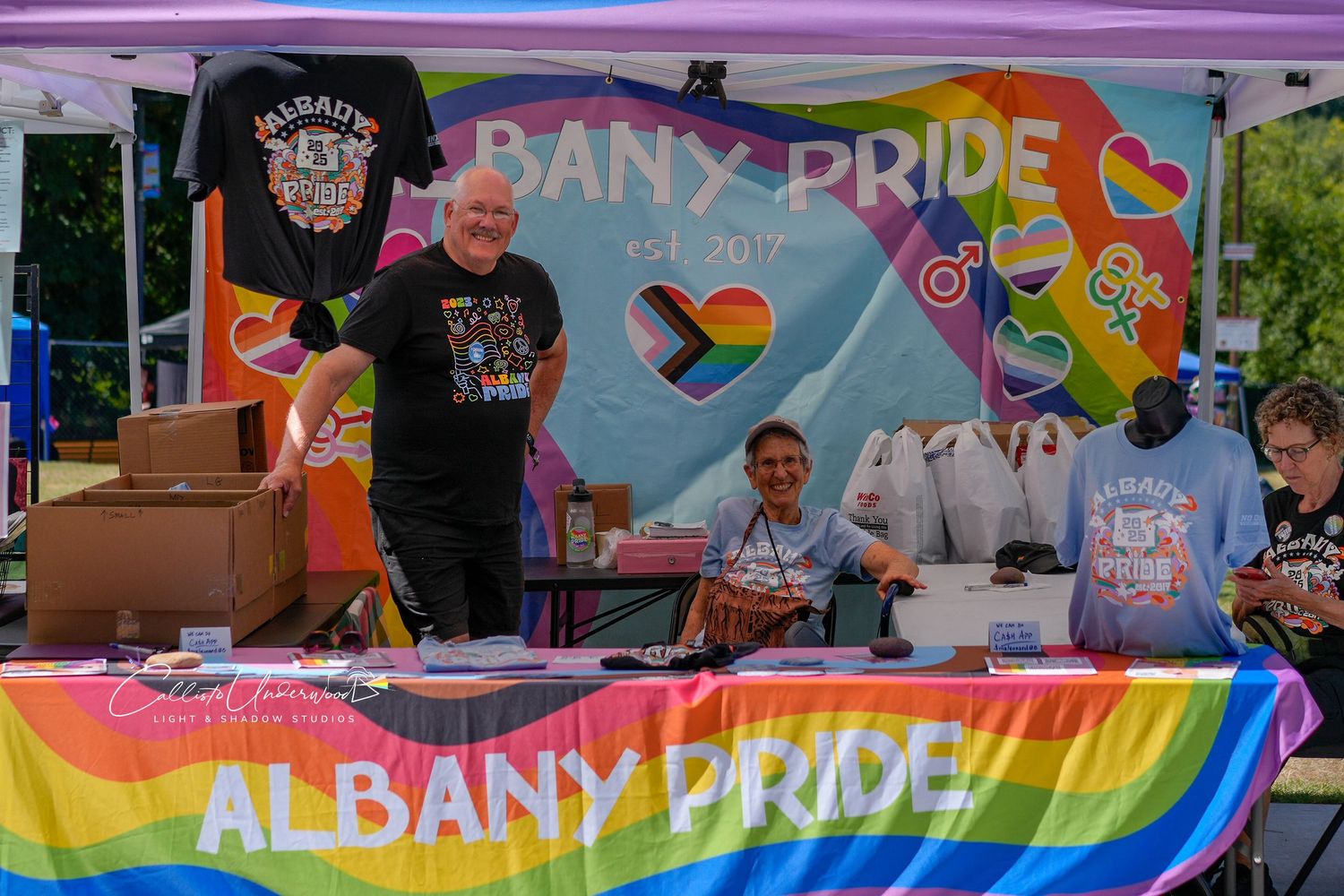 Smiling volunteers at the Albany Pride booth display t-shirts and merchandise on a rainbow tablecloth at an outdoor festival.