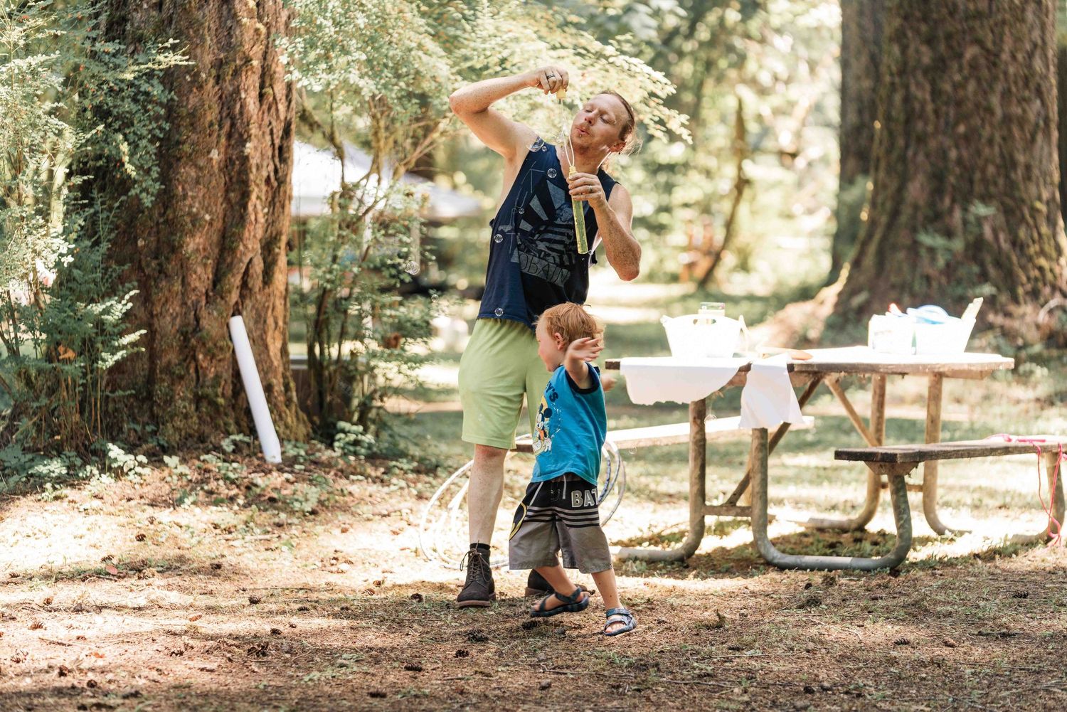 A woman in a green skirt lifts a toy overhead while a small boy in a blue shirt reaches up near a picnic table in the forest.