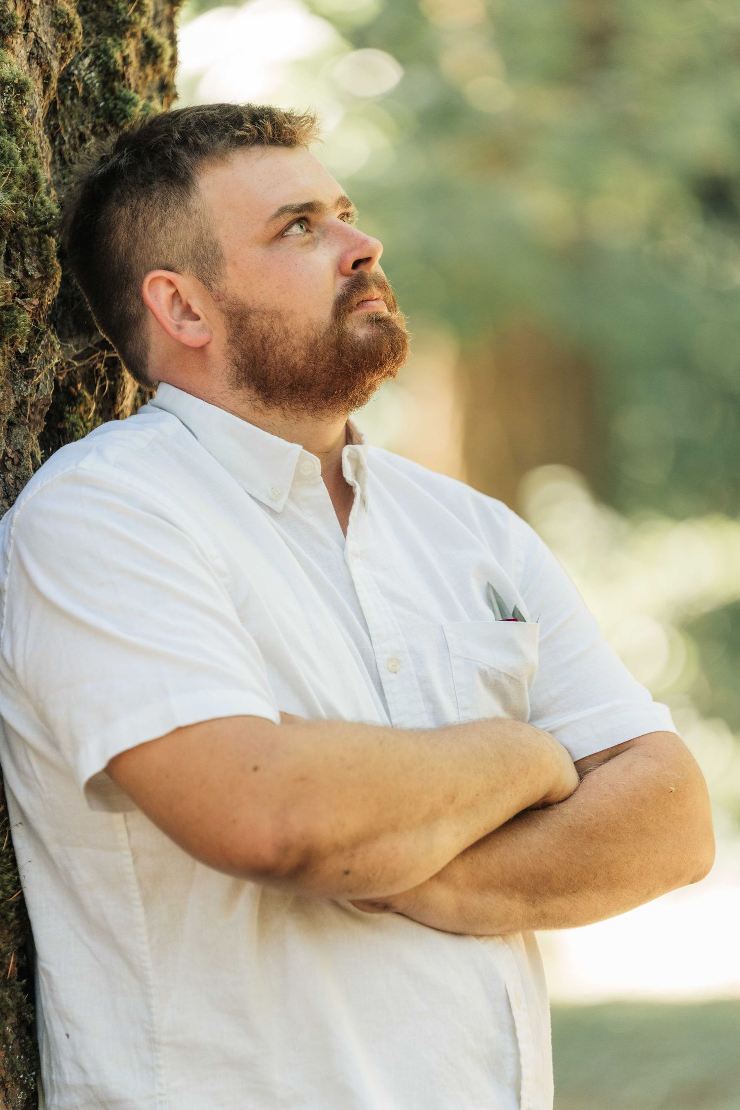 A bearded man in a white shirt crosses his arms and gazes upward while leaning against a mossy tree trunk.