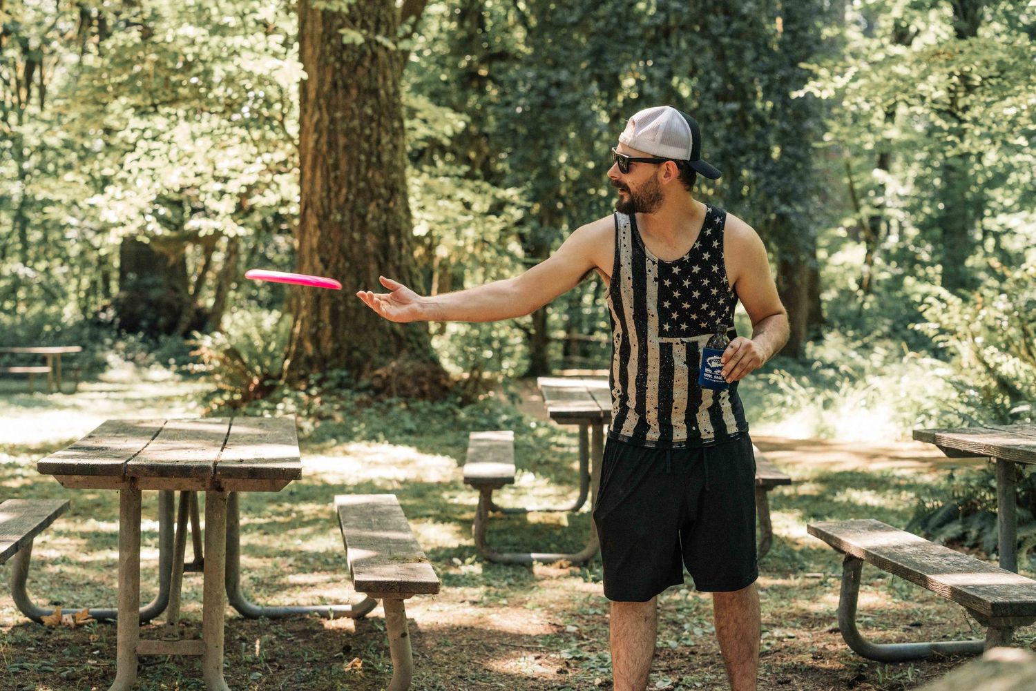 Man in a flag tank top and cap tosses a pink frisbee near wooden picnic tables in a shaded forest campground.