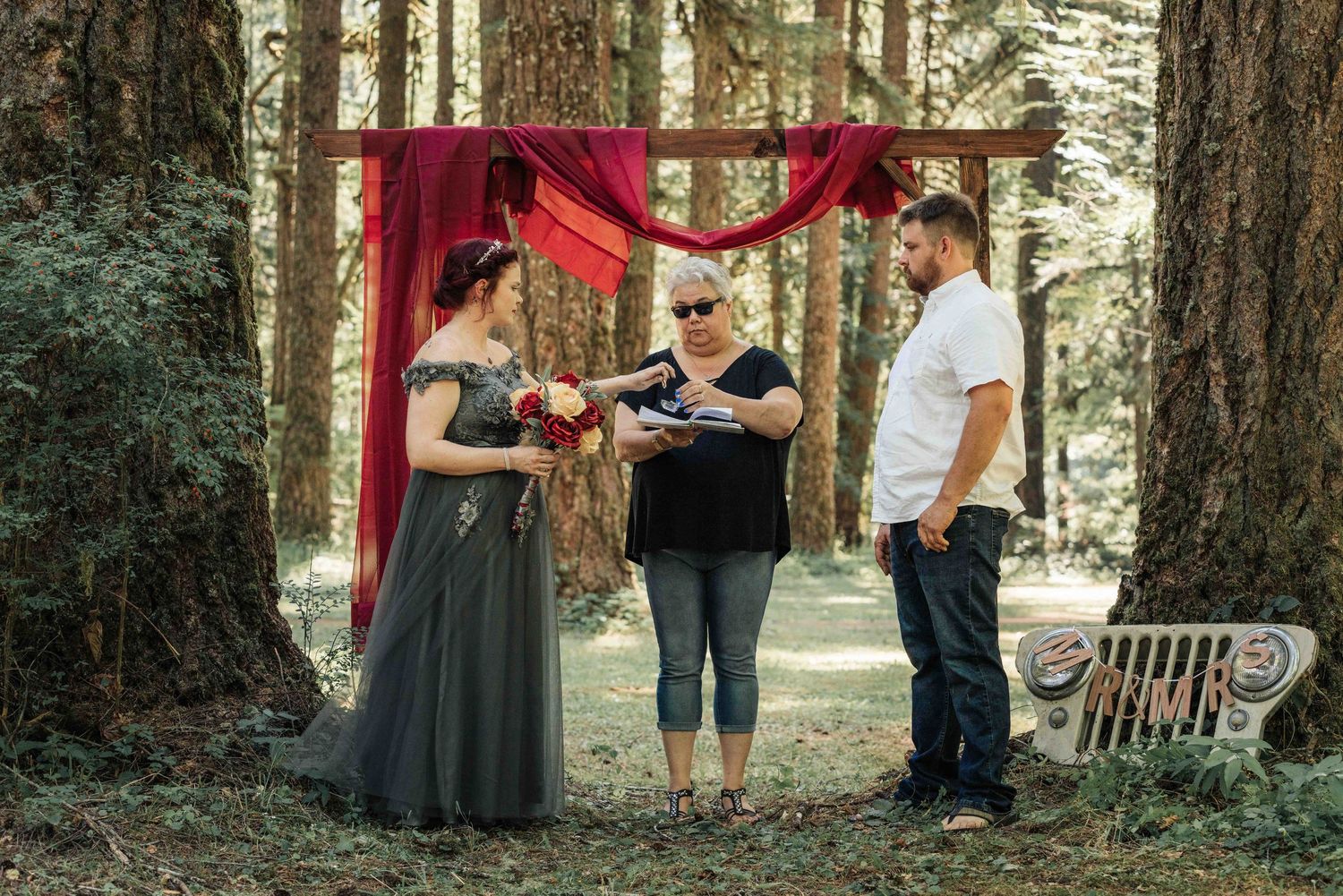 A wedding ceremony in a redwood forest with a bride, groom and officiant standing beneath a rustic wooden arch with crimson drapery.