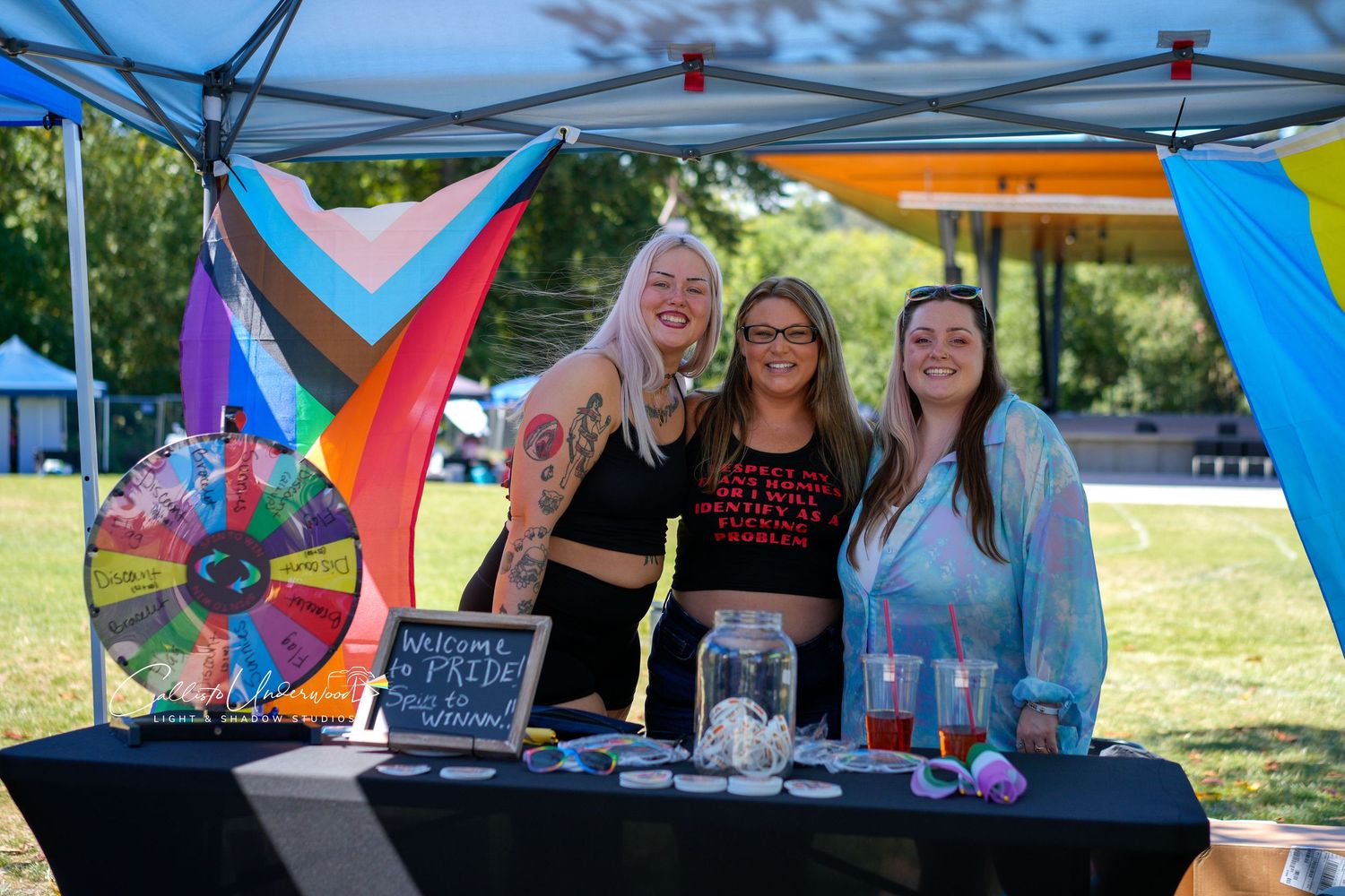Three smiling women stand behind a table at a vendor booth with a prize wheel, drinks and a rainbow pride flag draped on the canopy.