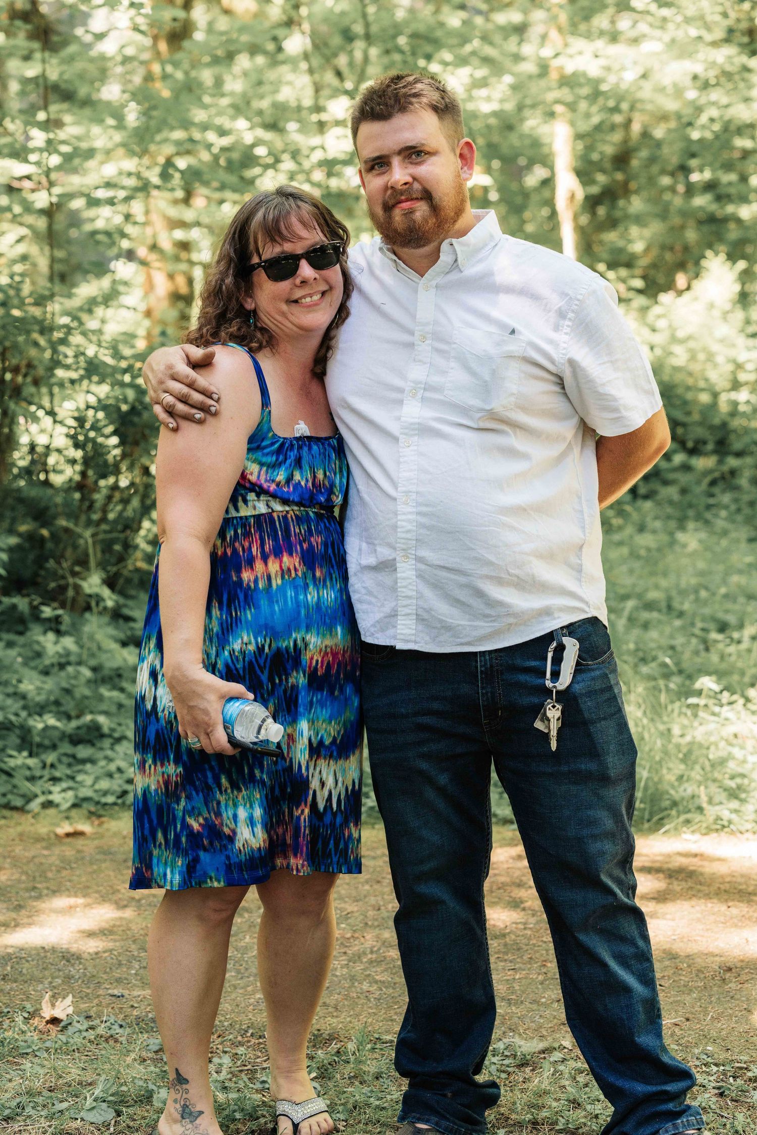 Smiling couple posing arm in arm outdoors in a sunlit forest, she in a colorful dress and sunglasses, he in a white shirt.
