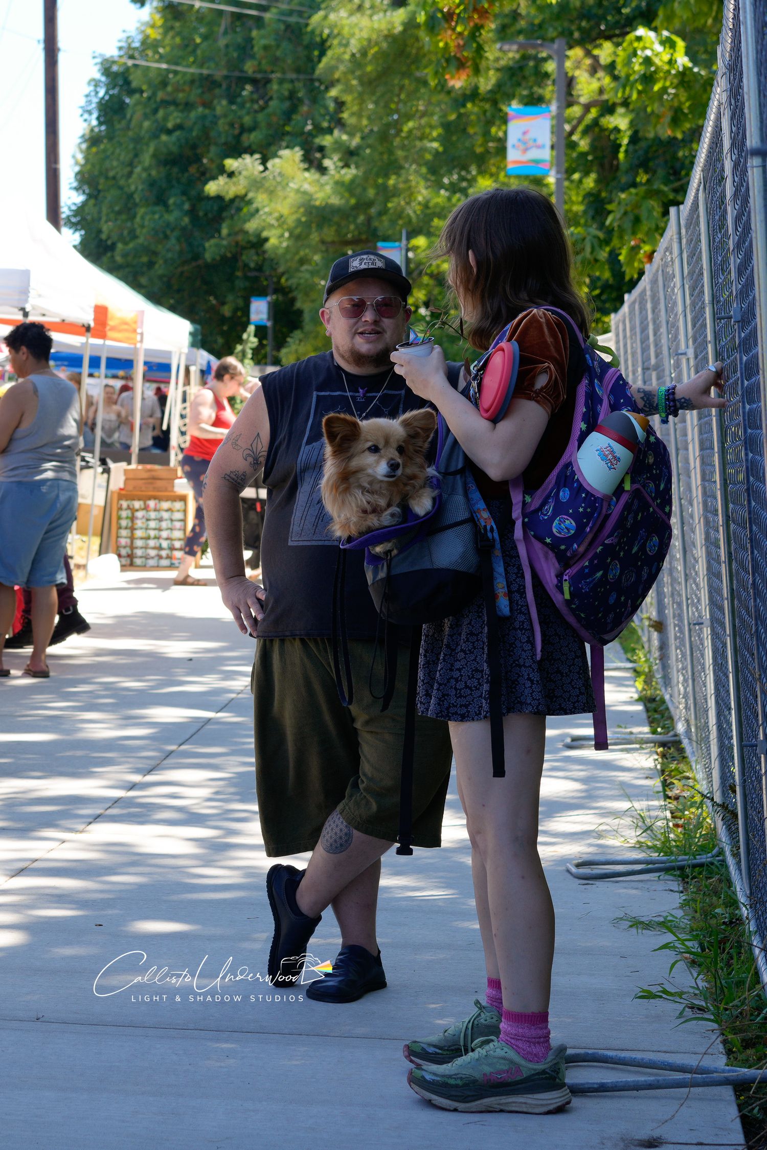 Man in a black cap carries a small fluffy dog in a front pouch while talking with a young woman in a purple backpack at a street fair.