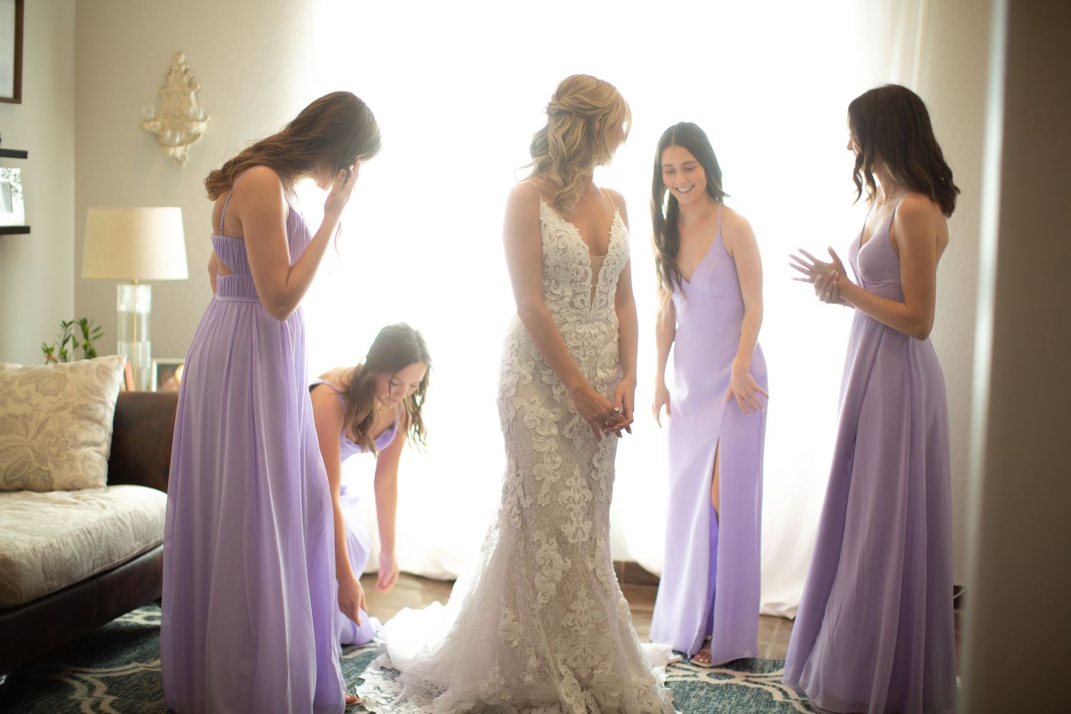 Wedding party in purple dresses and white gown gather by window during preparations.