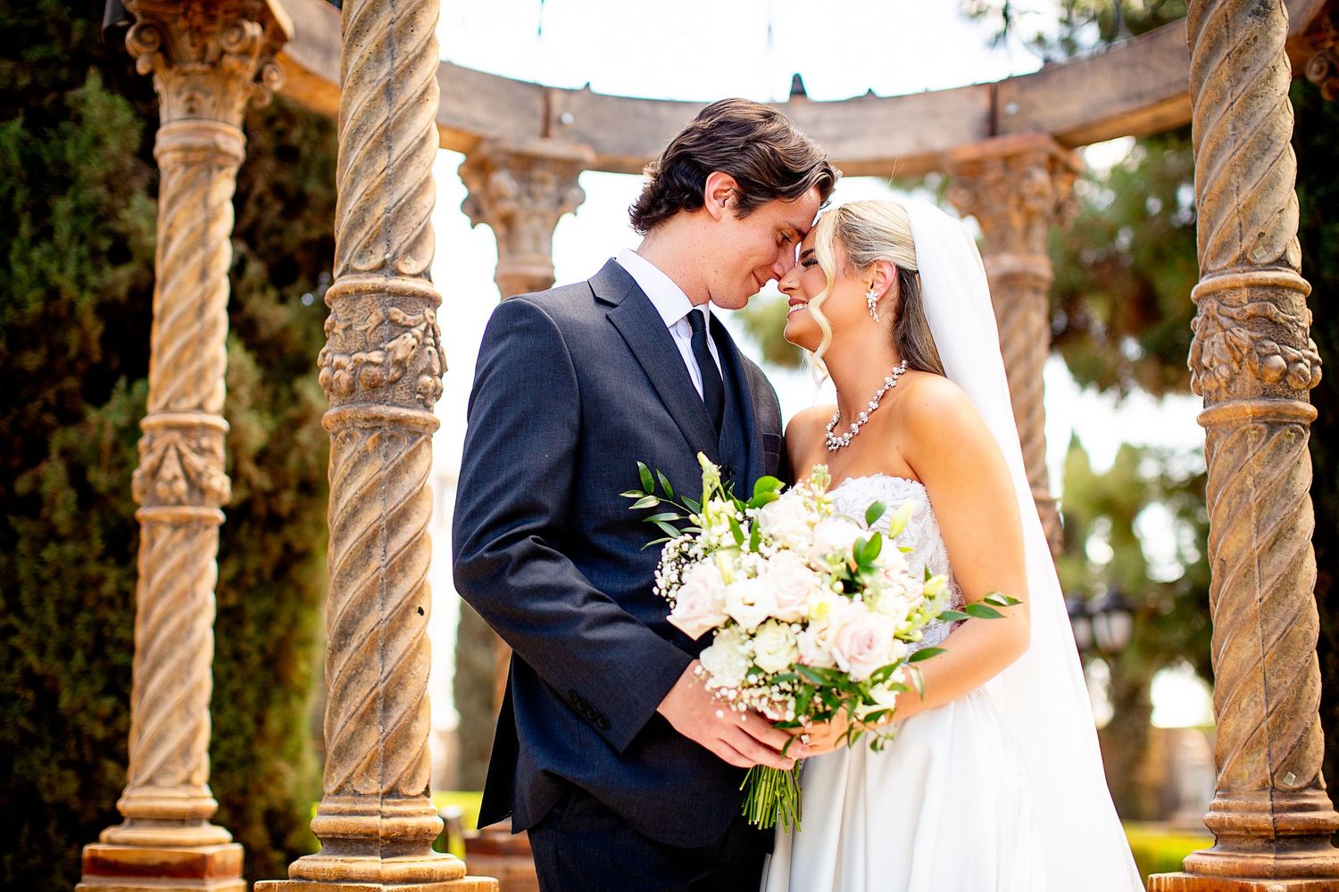 Wedding couple posing between elegant stone pillars in an outdoor garden setting.
