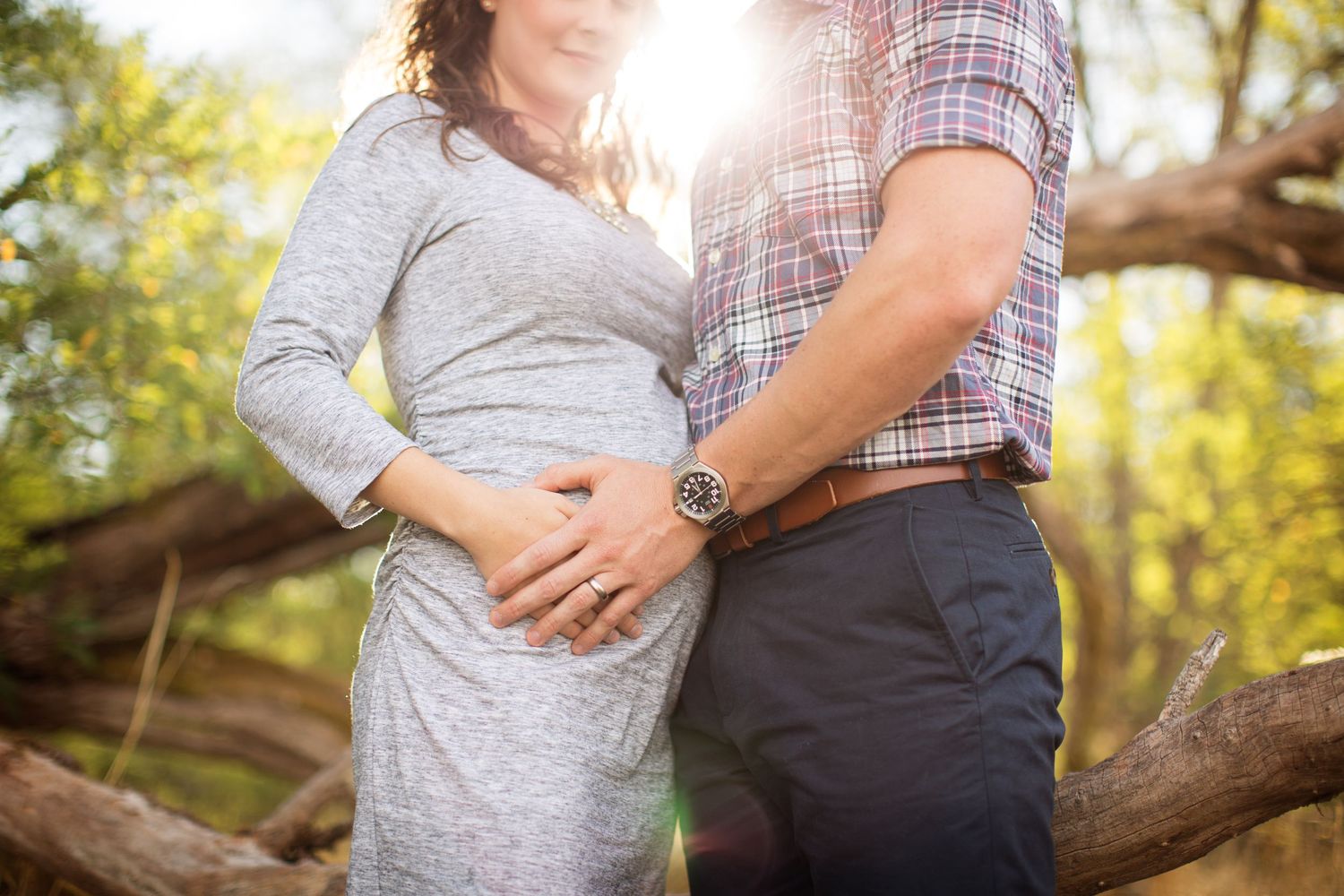 Close up of expectant couple holding hands in sunlit forest setting.