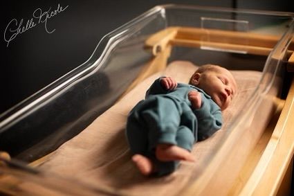 A small baby wearing blue clothing rests on a wooden surface in natural light.