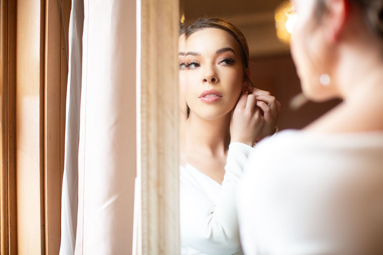 A person in white adjusts their earring while looking in a mirror by a window.