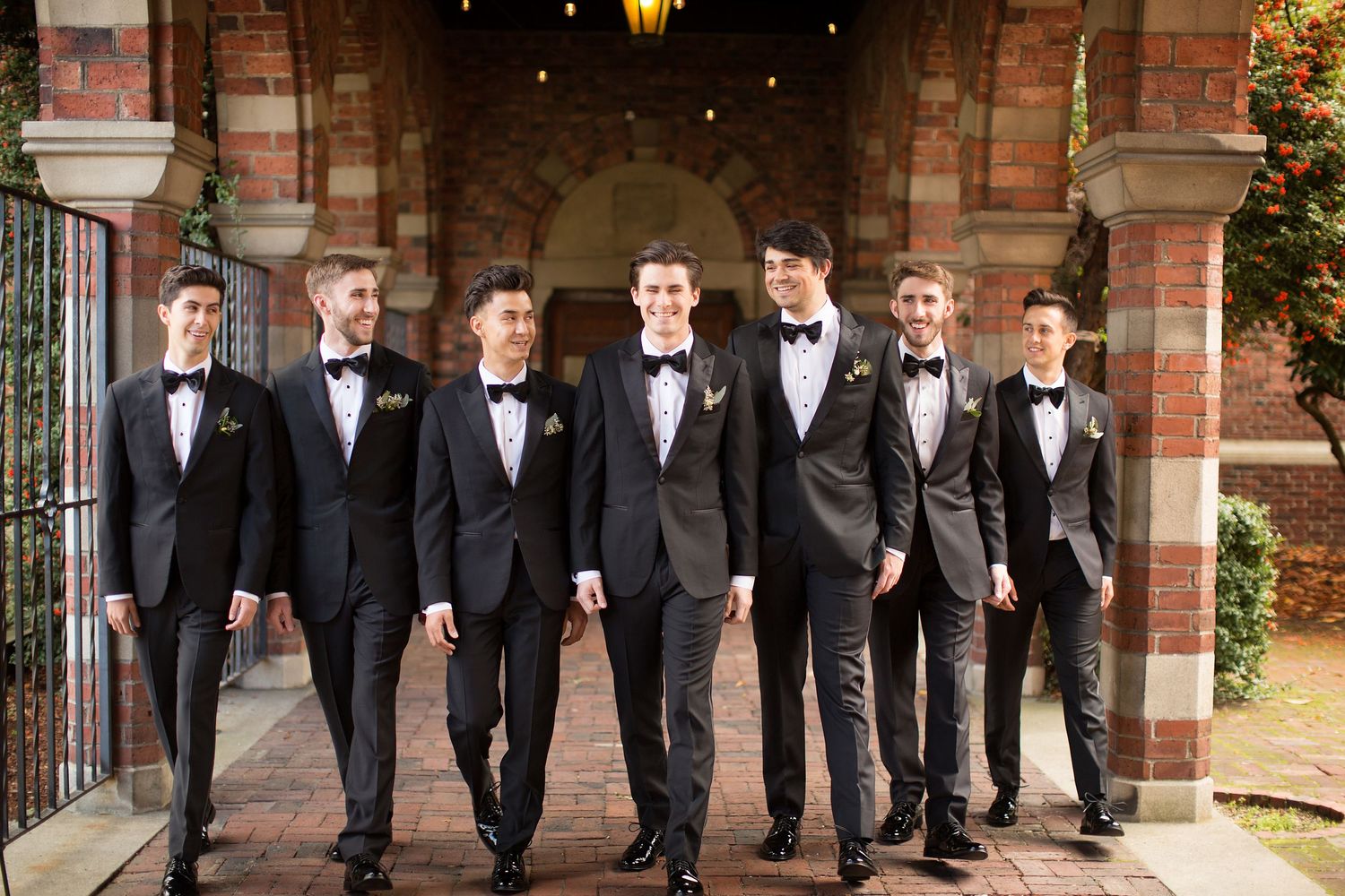 Groomsmen in black tuxedos pose together against a brick building backdrop.