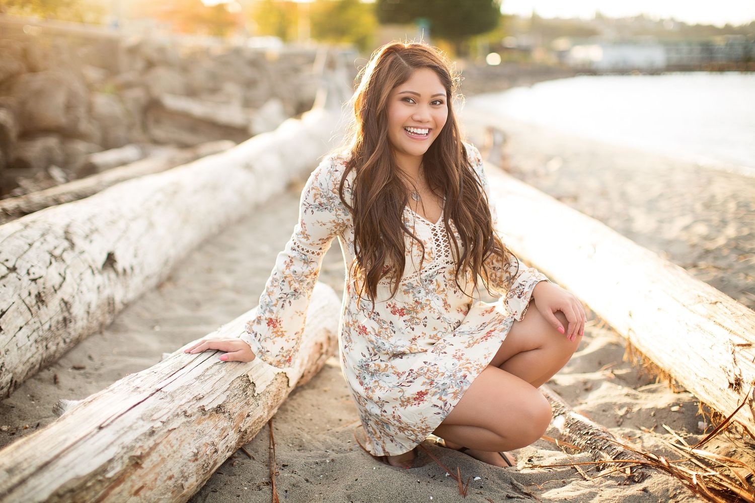 Woman in floral dress poses on driftwood at beach during golden hour.
