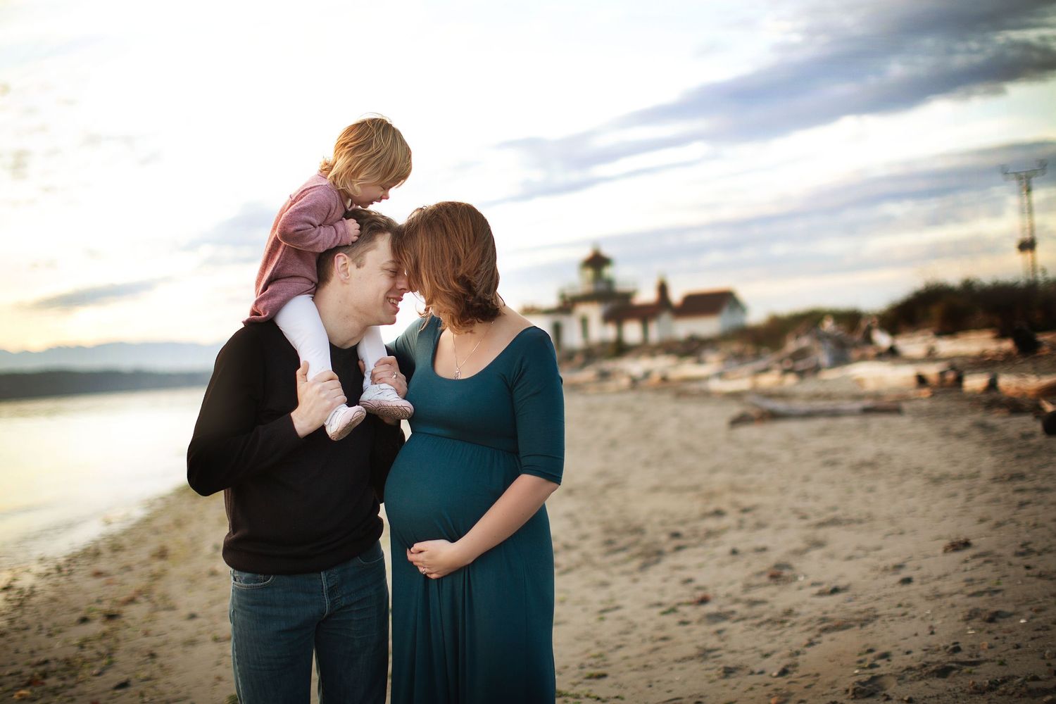 Family shares intimate moment on beach at sunset with lighthouse in background.