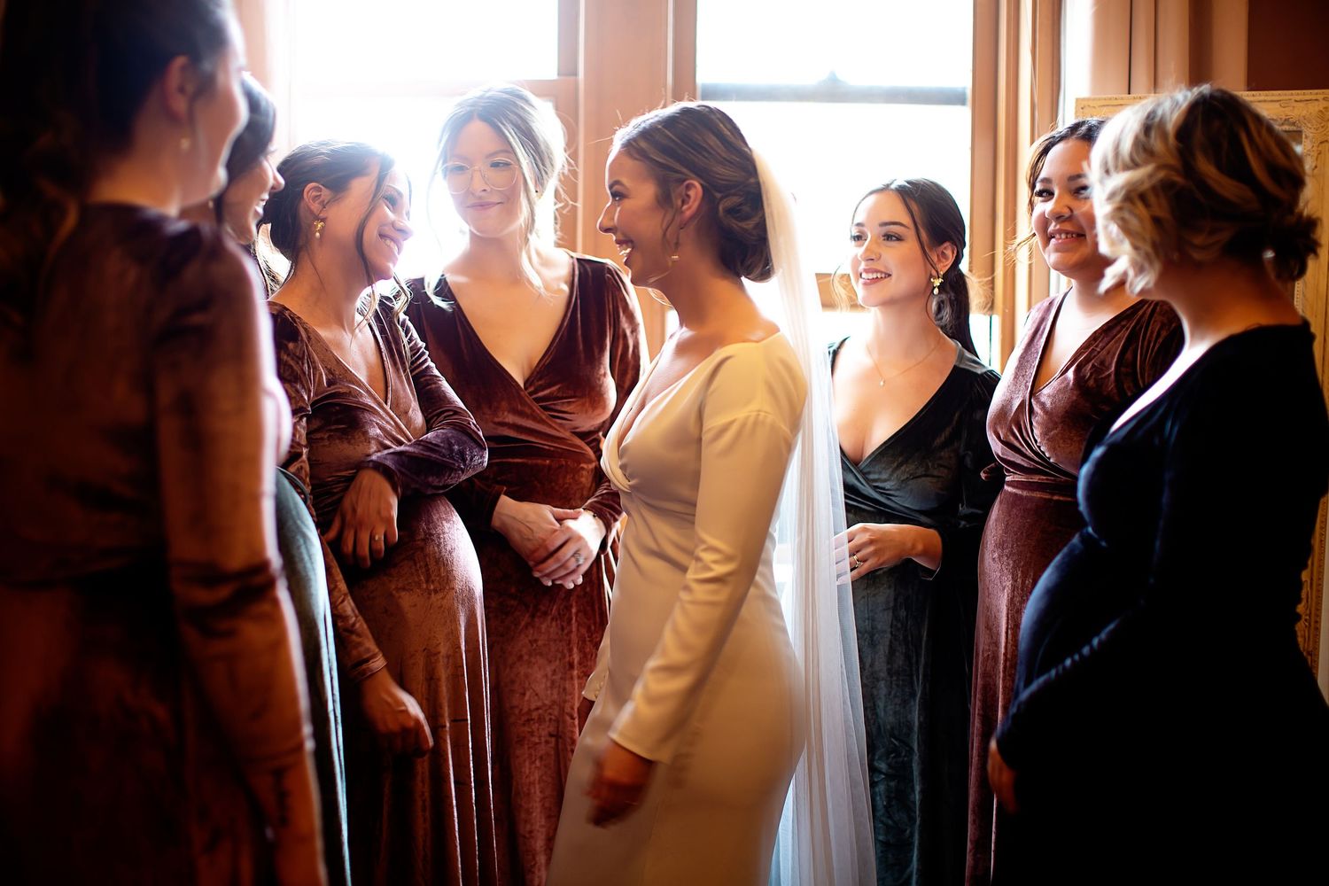 A group wearing burgundy and dark dresses gather in warm window light.