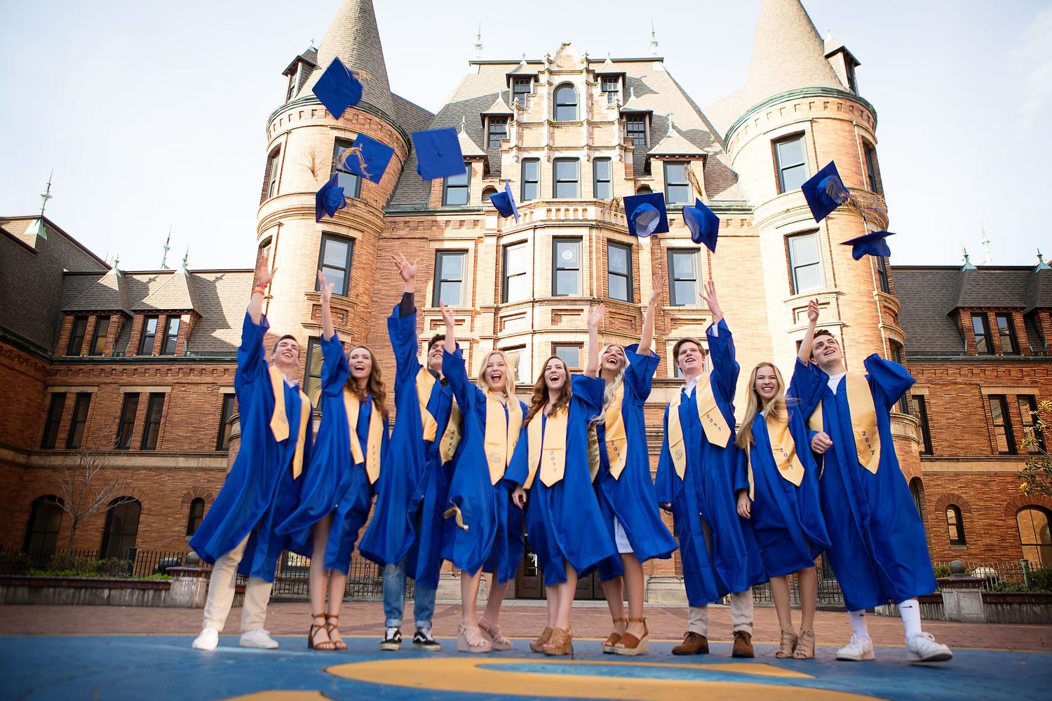 Group photo of graduates in blue gowns throwing caps in front of historic brick building.