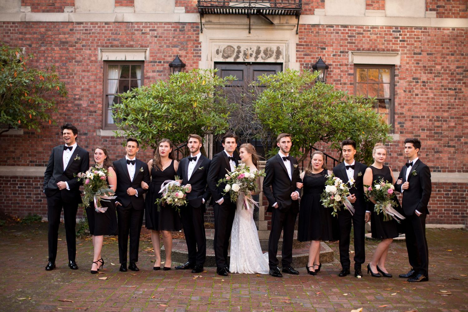 Wedding party members gather for formal photos outside a historic brick building.