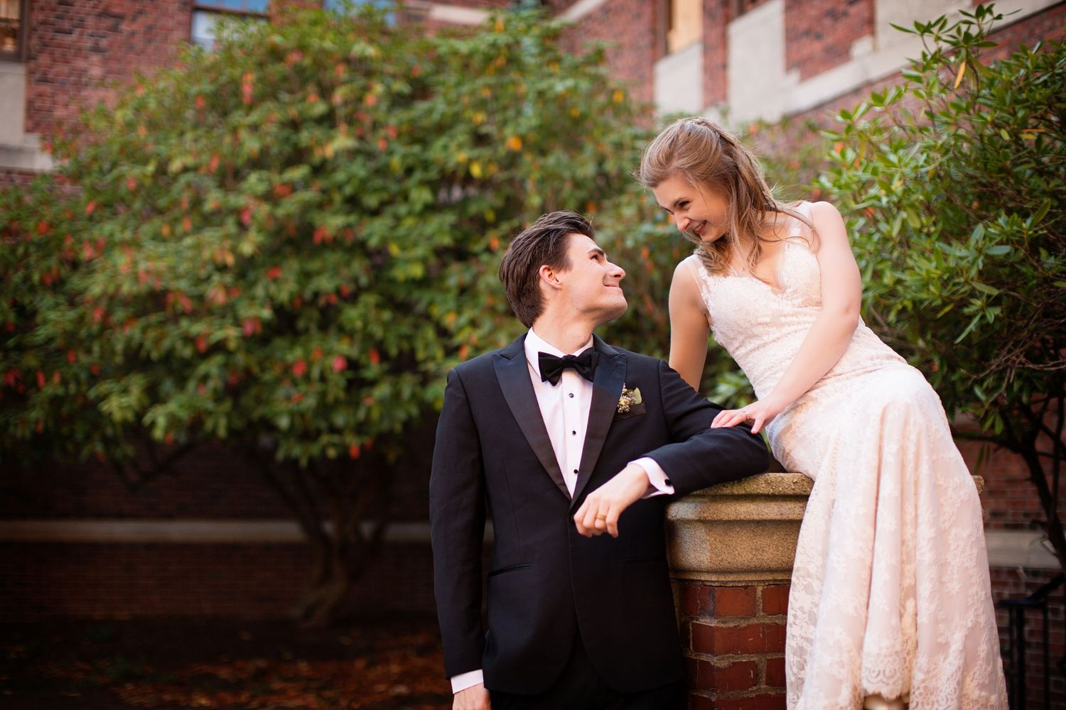 Wedding couple poses among lush greenery against brick building backdrop.