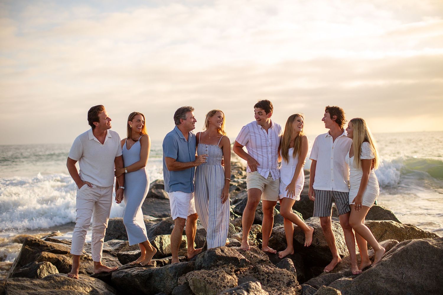 A group in white and blue clothing sits together on coastal rocks at sunset near the ocean.