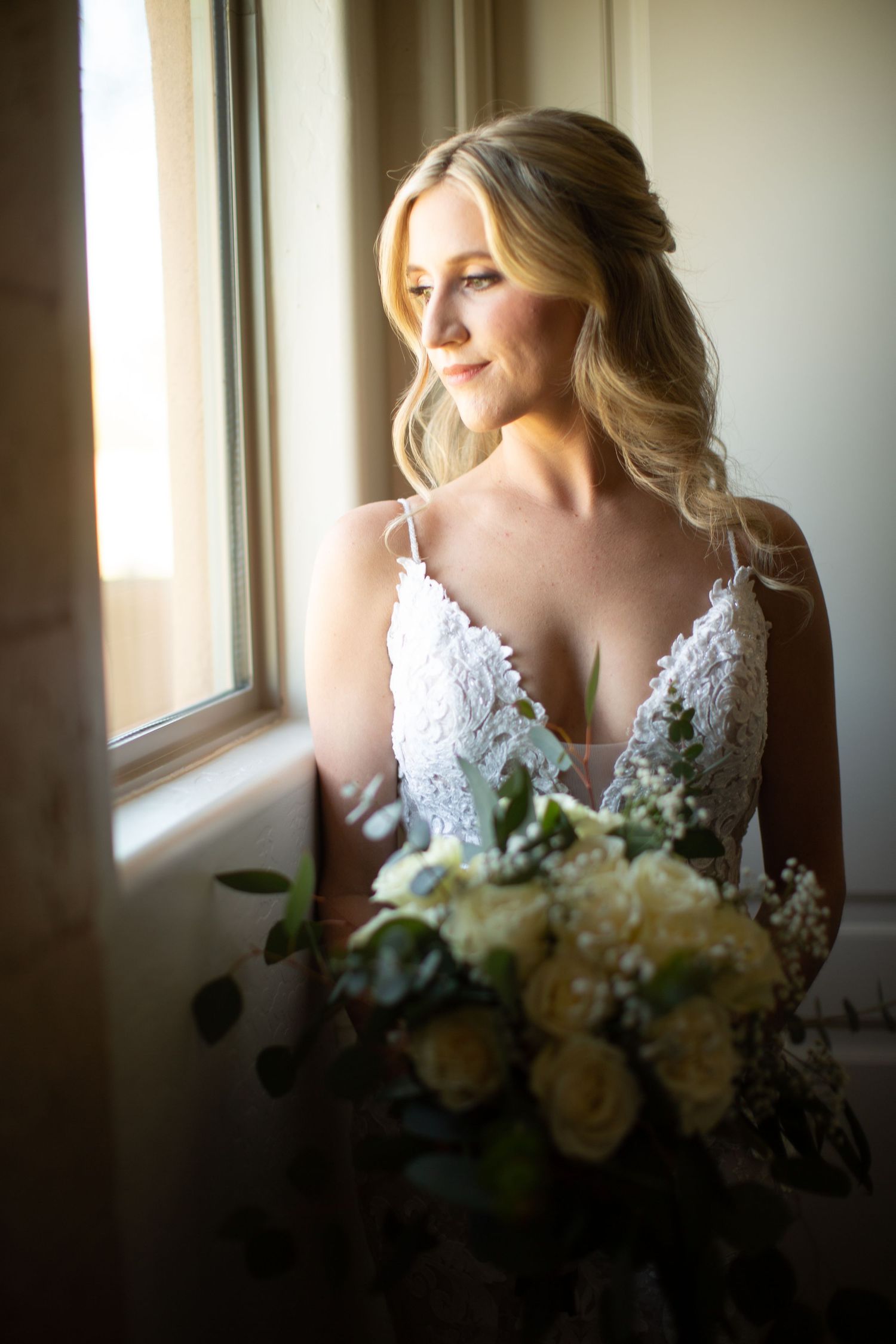 Bride in lace wedding dress poses by window with white floral bouquet in soft natural light.