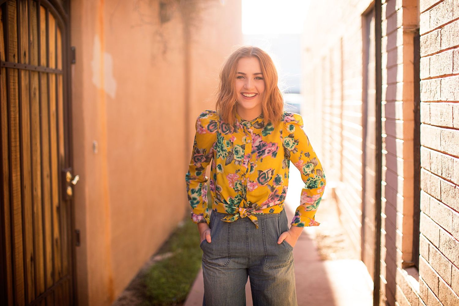 Woman in yellow floral top poses against brick wall in natural lighting.