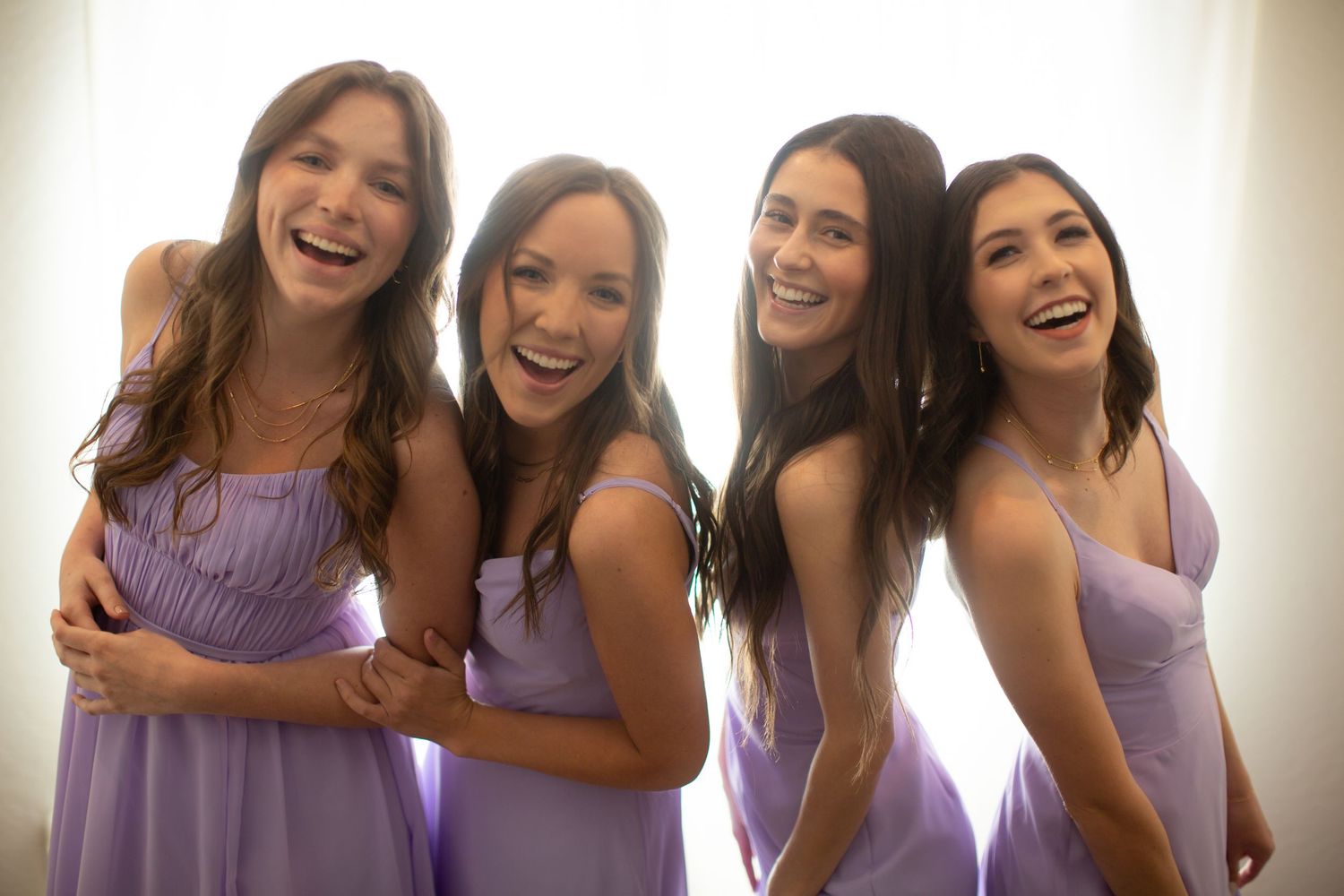 Bridesmaids in light purple dresses laugh and smile together during pre-wedding celebrations.