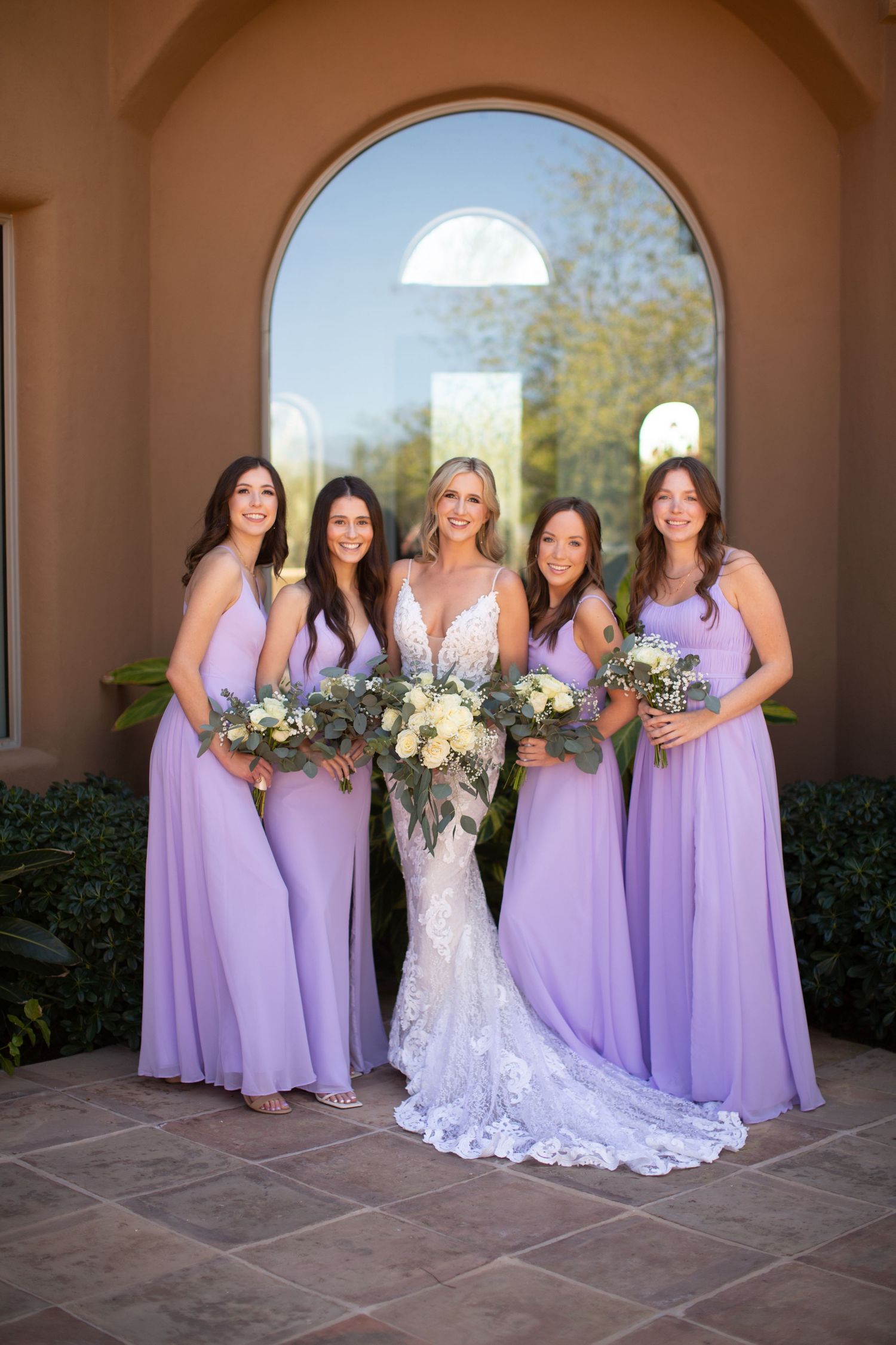 Wedding party poses in archway with bridesmaids in lavender dresses alongside bride in white lace gown.