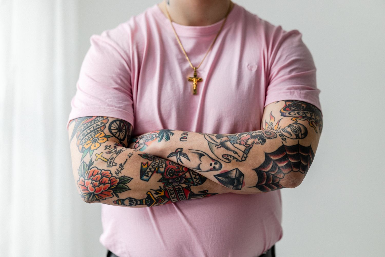 Yksityiskohtakuva miehen torsosta. Miehellä on päällä pinkki t-paita ja iso risti kaulassa. Käsivarret on tatuoitu täyteen värikkäitä kuvia.