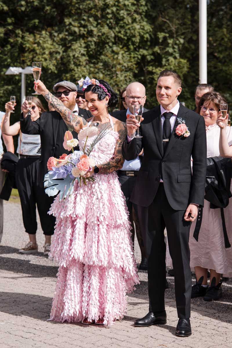 Hääpari skoolaa skumppalasien kanssa vieraiden kanssa. Vieraita näkyy taustalla. Tervetulopuhe on juuri pidetty.