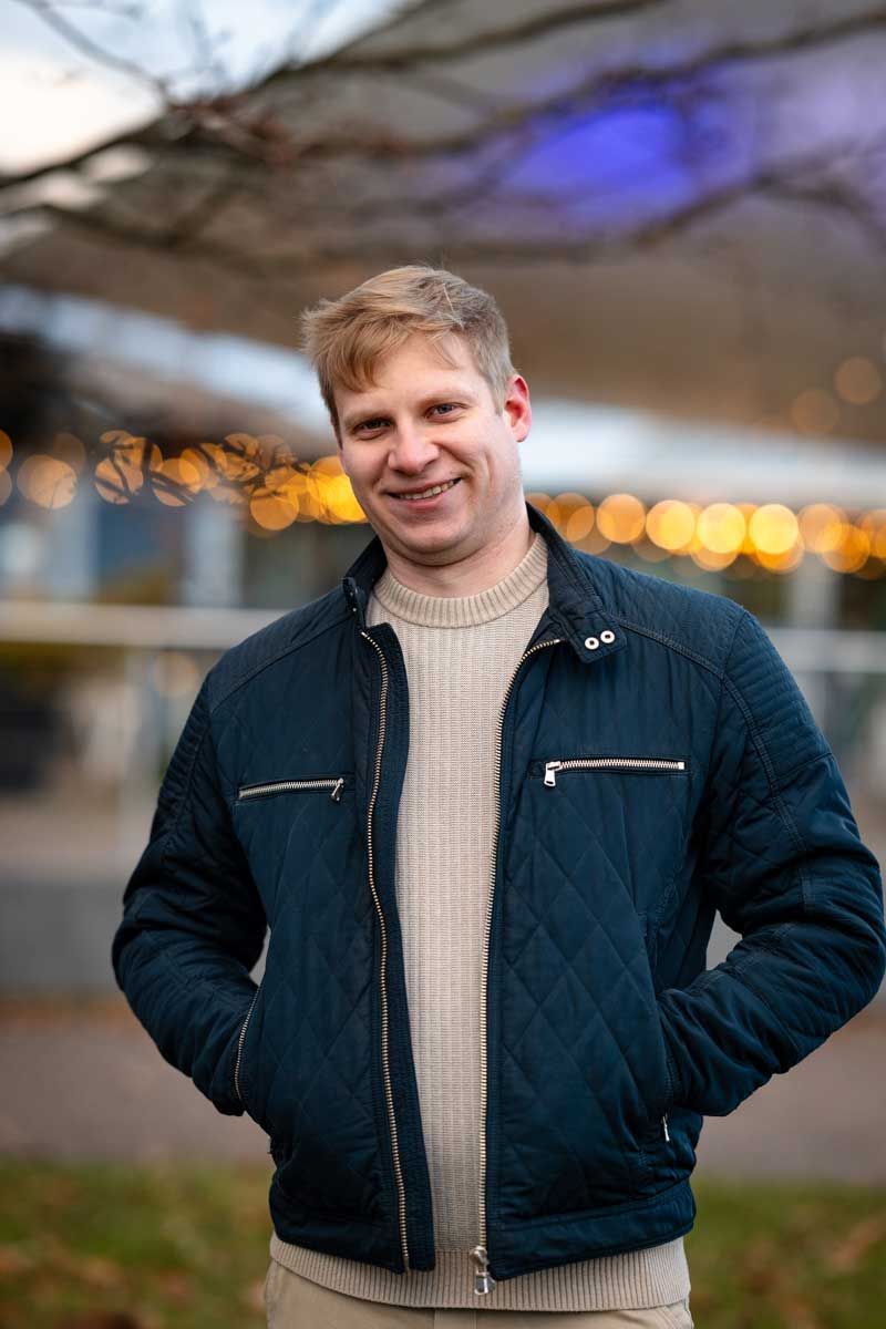 Mies seisoo takki päällä kädet taskussa kahvilan edessä. Kahvilan valot jää kauniisti blurriksi taustalle. Mies katsoo kameraa ja hymyilee.