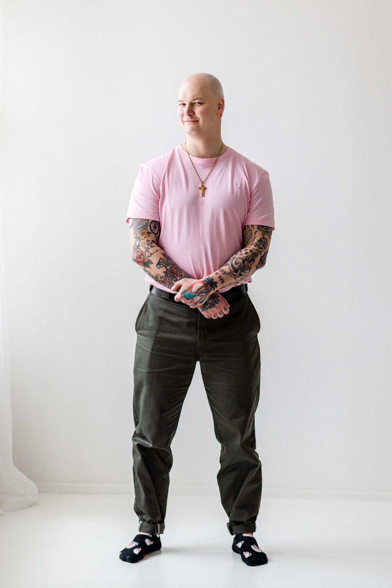 Tatuoitu kalju mies seisoo. Miehellä on pinkki t-paita päällä ja tumman vihreät housut. Mies seisoo vaaleassa studiotilassa.