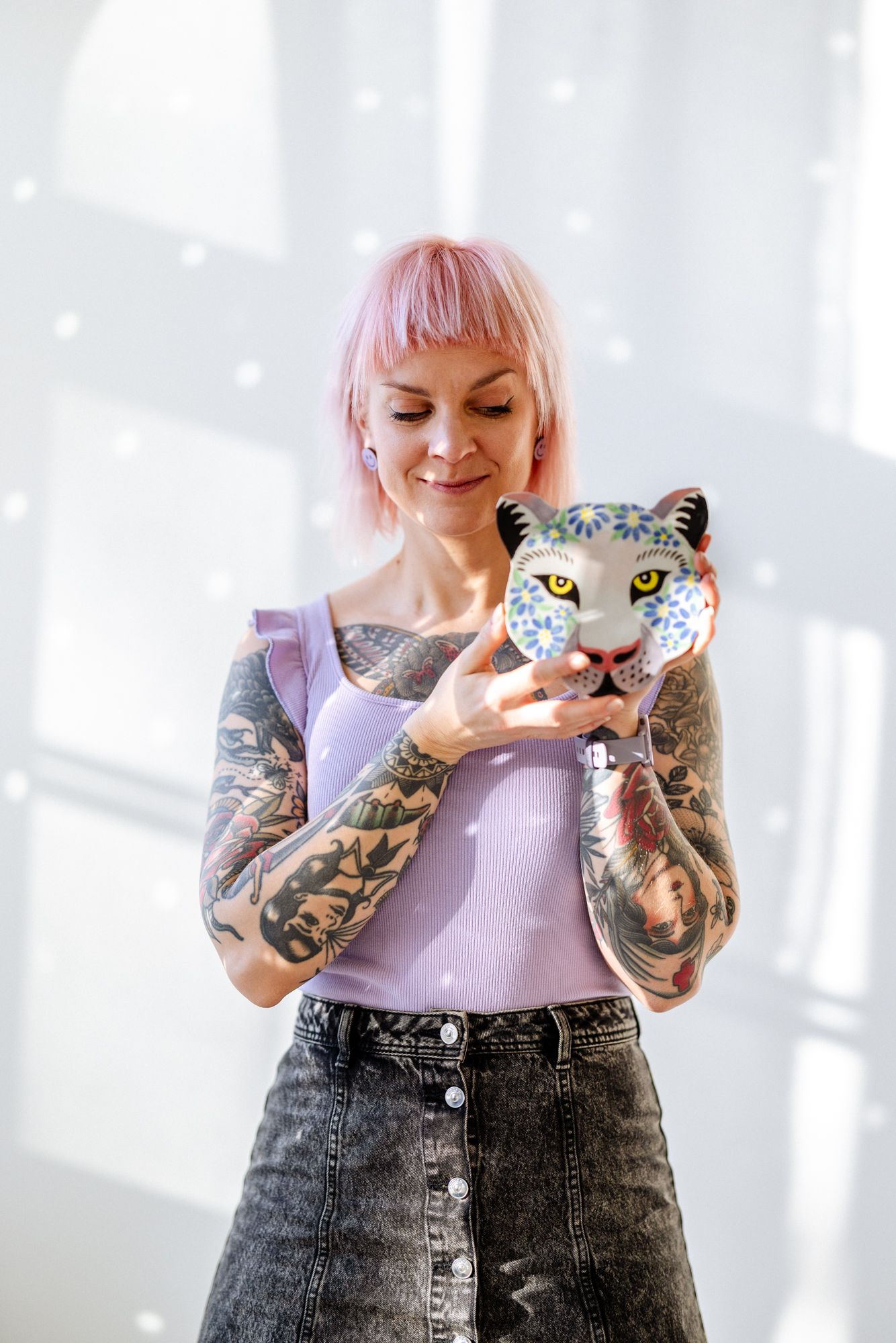 Tatuoitu nainen esittelee omatekemäänsä keraamista pantterinpää seinäkoristetta. Naisella on pinkit hiukset. Tila on valoisa ja valkoinen.
