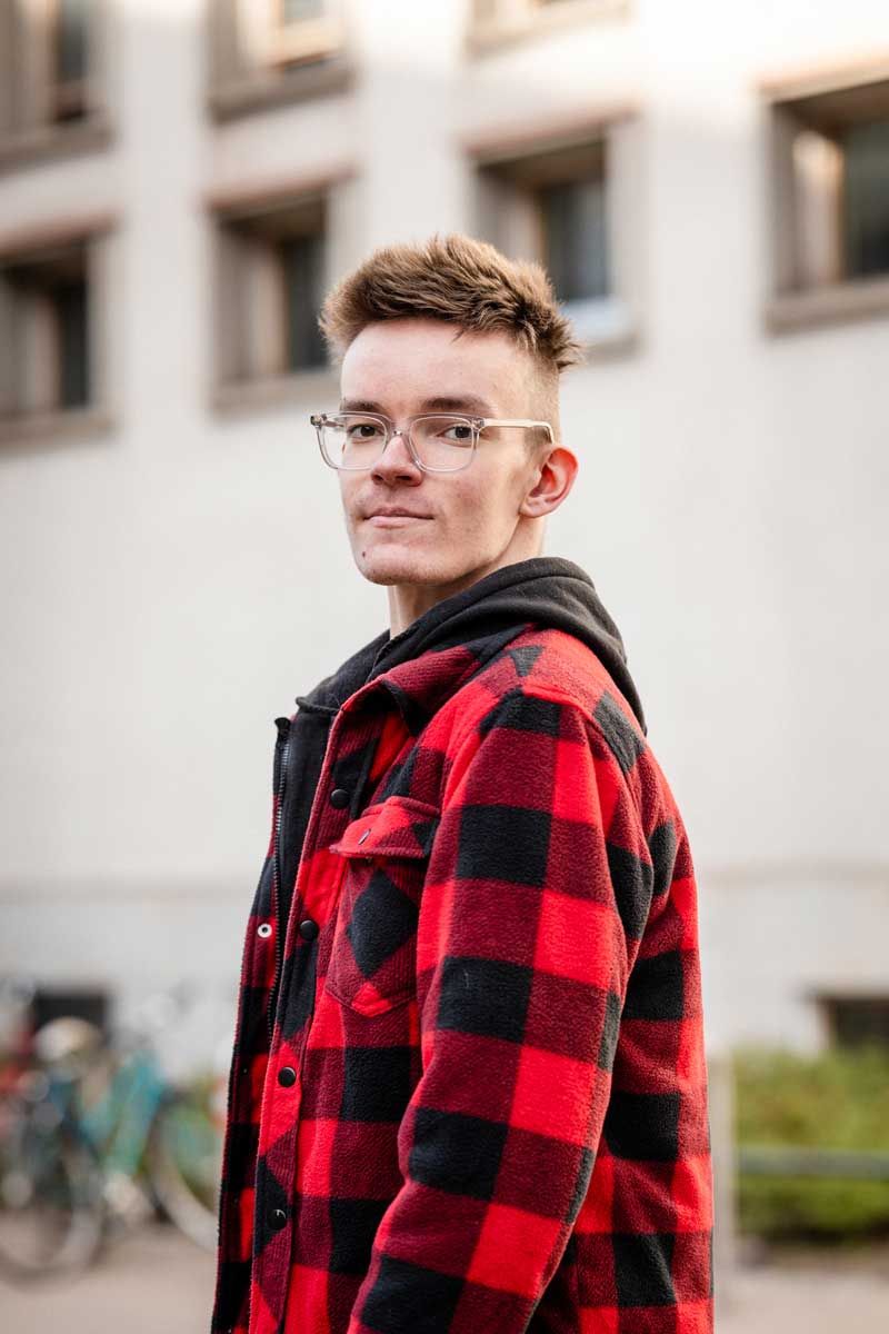 Nuori mies kävelee kadulla Helsingissä ja katsoo olan yli suoraan kameraan. Miehellä on silmälasit ja punamusta ruudullinen syystakki.