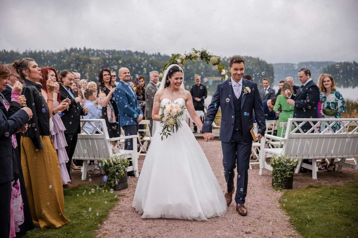 Hääpari vihkimisen jälkeen kävelemässä pois päin alttarilta. Vieraat puhaltaa saippuakuplia. Vihkiseremonia tapahtui järven rannalla.