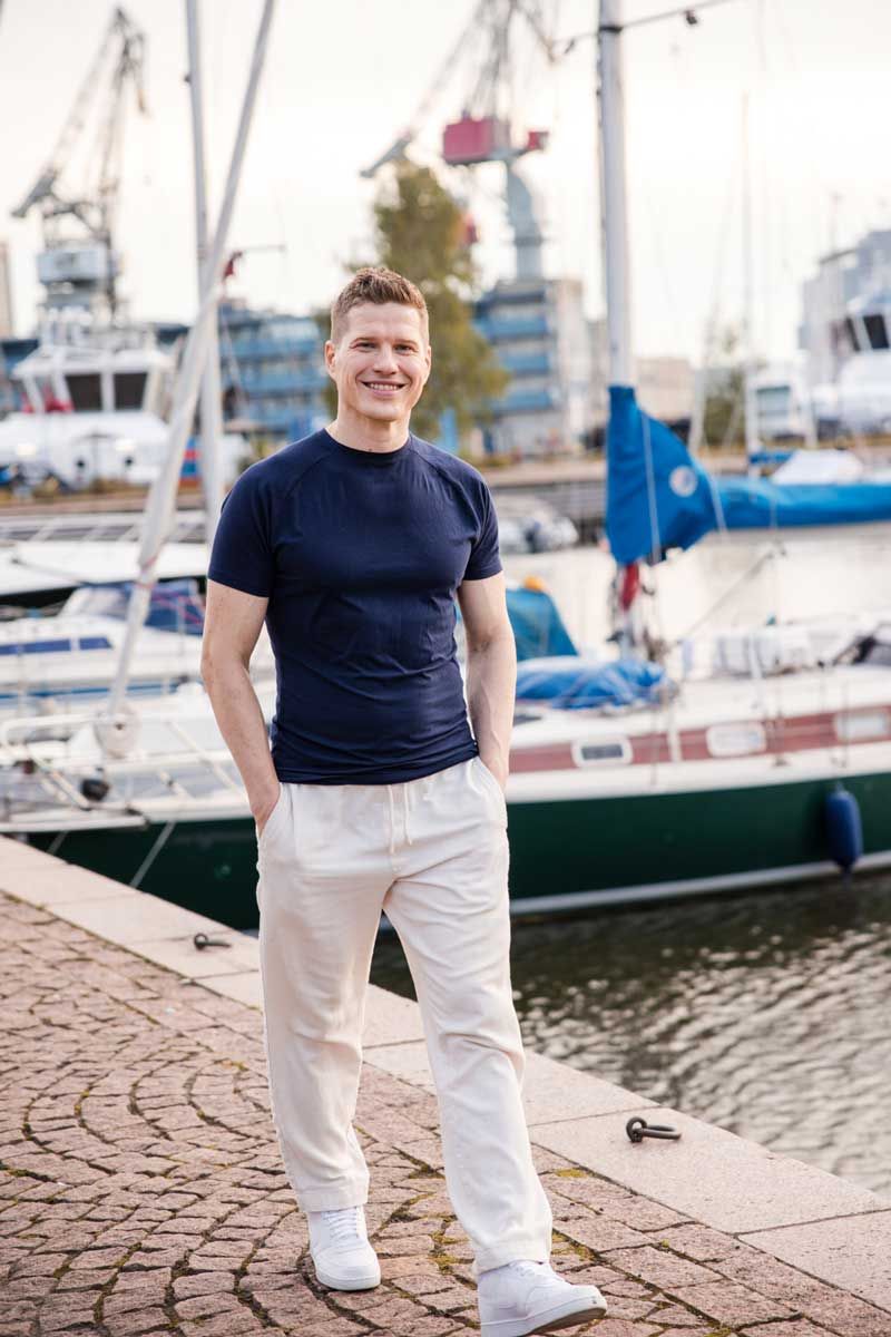 Komea mies kävelee rennosti satamassa tumma t-paita ja vaaleat pellavahousut päällä. Katse on kameraan ja hymy huulilla.
