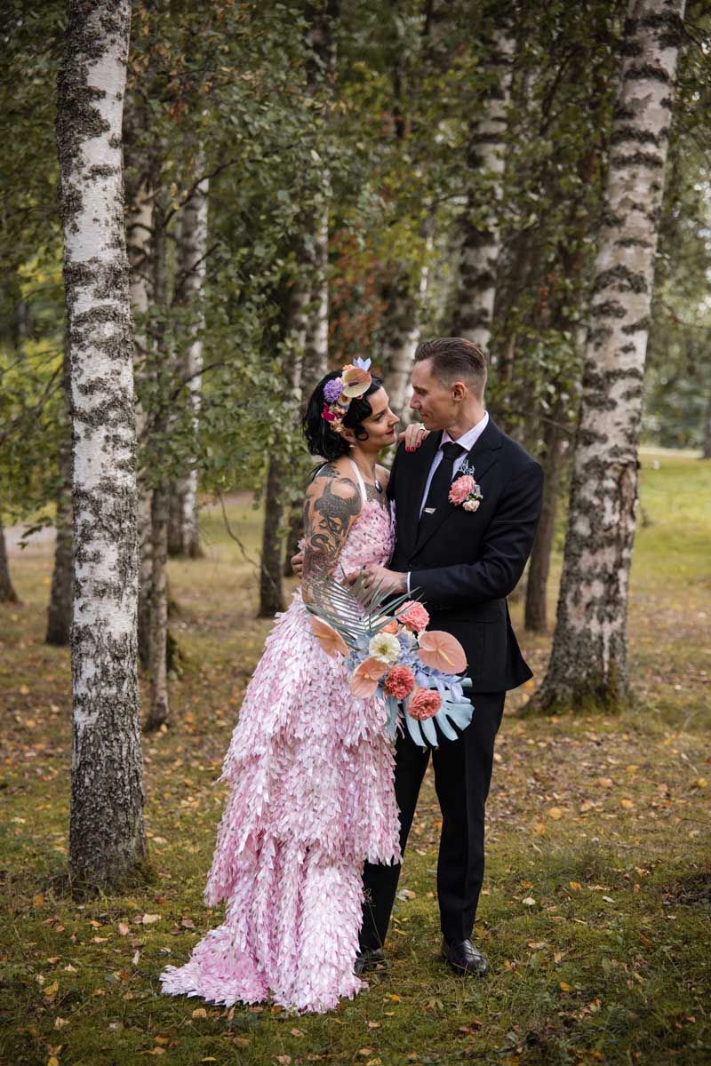 Hääpari nojaa toisiaan vasten koivujen katveessa. Morsiammella on pinkki kimalteleva hääpuku. Sulhasella on tumma puku ja pinkki viehe.