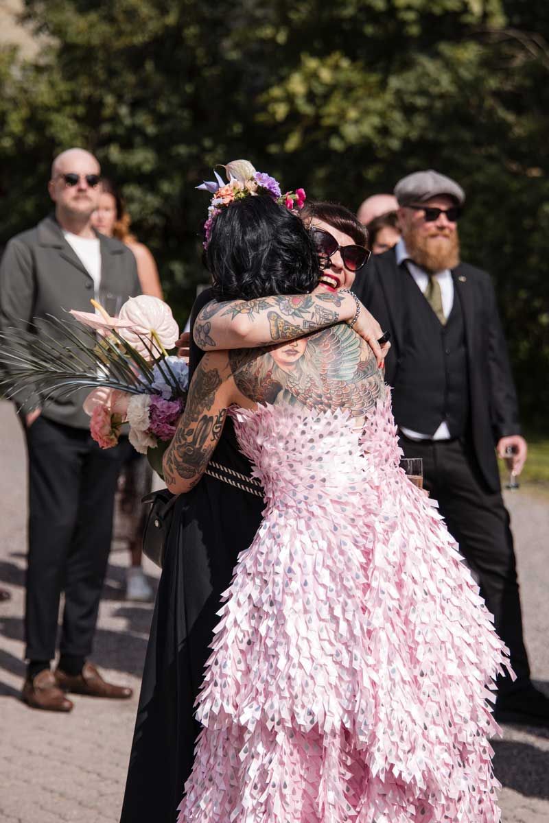 Morsian halaa juhlavierasta lämpimästi. Morsiammella on näyttävä pinkki juhlapuku päällä ja iso tatuointi selässä.