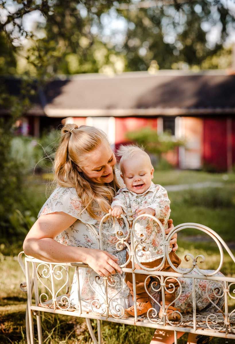 Äiti ja 1-vuotias omalla pihalla. Äiti istuu puutarhatuolissa ja taapero seisoo äidin vieressä ja äiti pitää kiinni ja katsoo lempeästi.