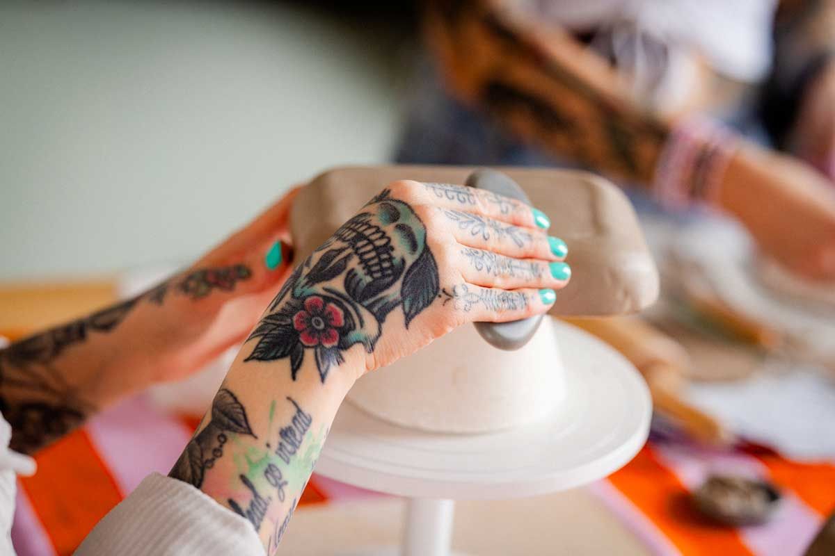 Yksityiskohtakuva tatuoidusta kädestä työstämässä keramiikkaa. Turkoosit kynnet. Kuvassa tasoitetaan keramiikka-astian pohjaa sileämmäksi.