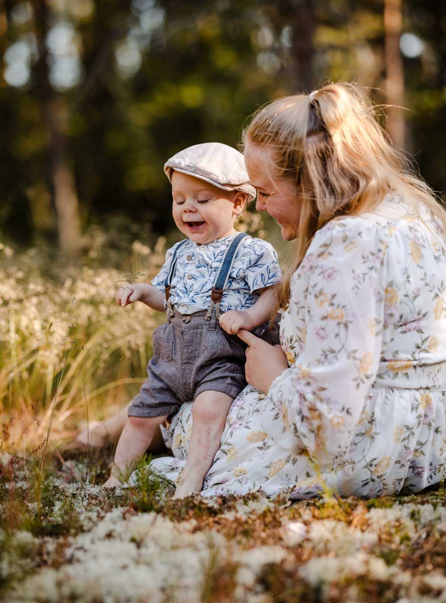 Äiti ja taapero istuu metsässä. Äiti pitää taaperoa sylissä ja molemmat on iloisia. 1-vuotias on puettu lakkiin ja henkselihousuihin.