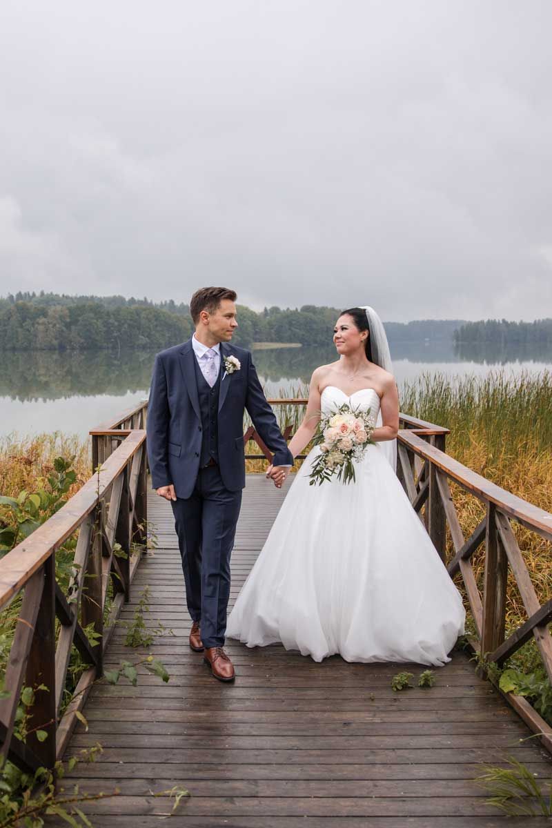 Hääpari kävelee laiturilla sumuisena ja harmaana päivänä. Pitävät käsistä kiinni ja katsovat toisiaan. Järvimaisema taustalla.