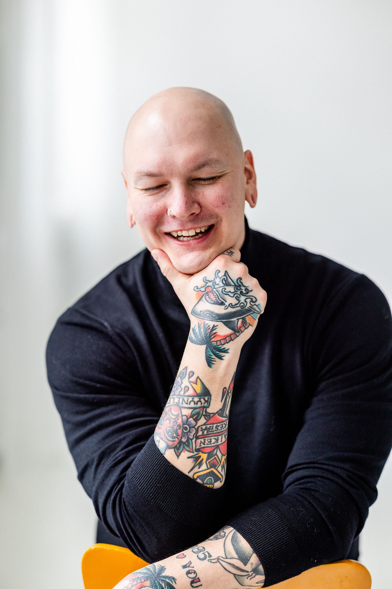 Tatuoitu kalju mies nojaa päätä kättä vasten. Miehellä on silmät kiinni ja hän nauraa. Kuva on otettu vaaleassa studiotilassa.