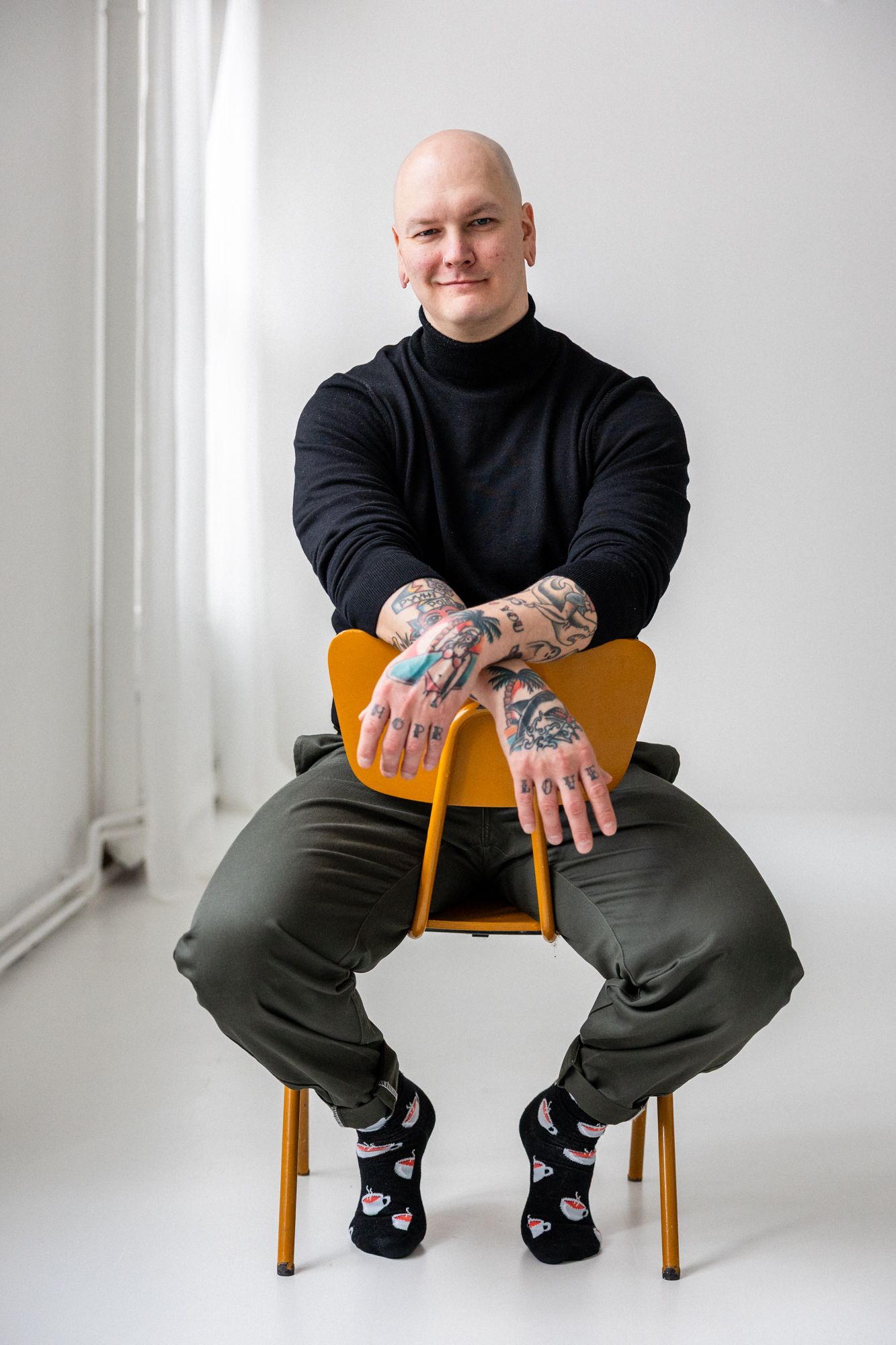 Tatuoitu kalju mies istuu tuolilla mikä on väärin päin. Miehellä on päällä musta polokauluksinen paita. Tila on vaalea ja avara.