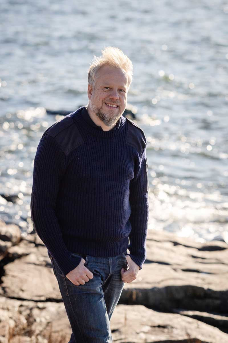 Mies kävelee rantakalliolla villapaita päällä. Meri aaltoilee taustalla. Aurinko osuu hiuksiin. Mies katsoo kameraa kohti ja hymyilee.