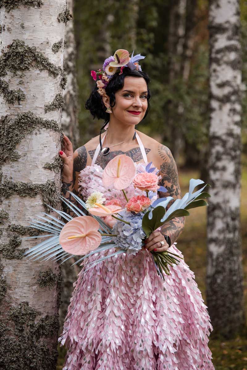 Morsian pinkissä hääpuvussa nojaa koivua vasten ja katsoo suoraan kameraan. Näyttävä ja persoonallinen kukkakimppu kädessä.