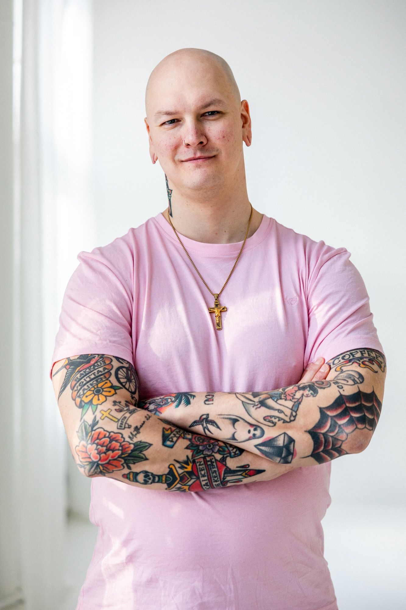 Tatuoitu kalju mies seisoo kameraa kohti. Miehellä on risti kaulassa ja pinkki t-paita päällä. Tatuoidut käsivarret on puuskassa.