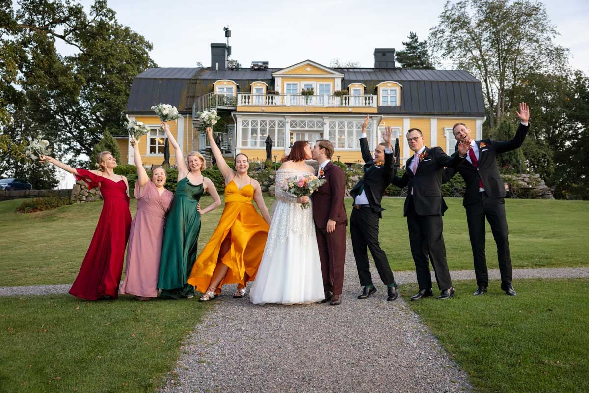 Ryhmäkuva hääparista kaasojen ja bestmanien kanssa Bodomin Kartanon pihalla Espoossa. Hääpari suutelee ja muut tuulettaa ja hurraa.