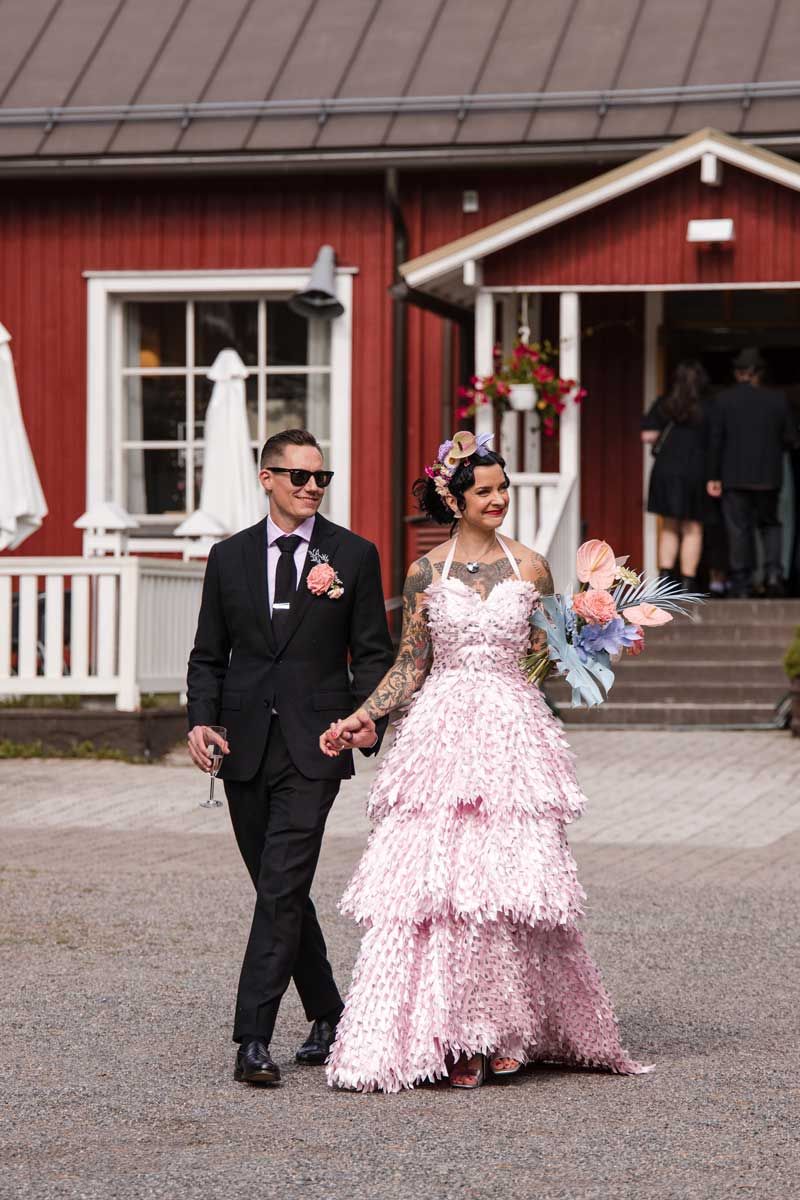 Näyttävä hääpari kävelee käsi kädessä juhlpaikan tilalla. Juhlapaikan punainen Hyrylän Torppa näkyy taustalla.